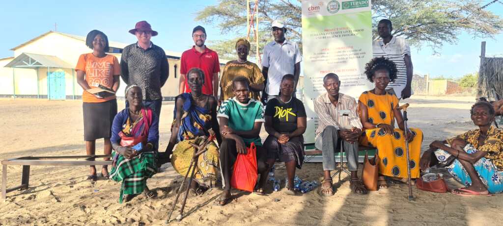 Members of the Nitiakara Disabled Self Help Group together with Dr. Manuel Rothe (standing, third from left), CBM Global Senior Cash and Livelihood Advisor, and Anthony Kimathi Ngari (standing, second from left), Humanitarian Coordinator of CBM Global Kenya. The group includes men and women of various ages, wearing colorful traditional and casual clothing. Some members are seated on benches, while others stand behind them, all smiling or looking serious at the camera. The background features a sandy ground and sparse vegetation.