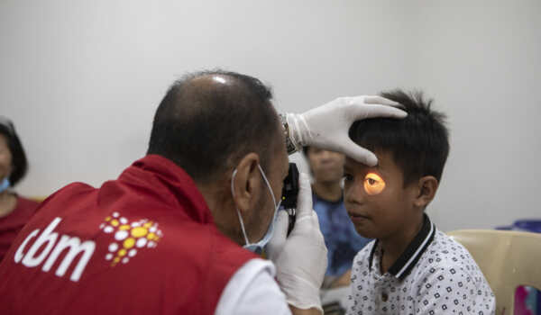 A healthcare professional in a red CBM vest performs an eye examination on a young boy using an ophthalmoscope. The boy sits still as the professional carefully inspects his eye, with a focused light shining on the eye. Other individuals are visible in the background, indicating a clinical setting.