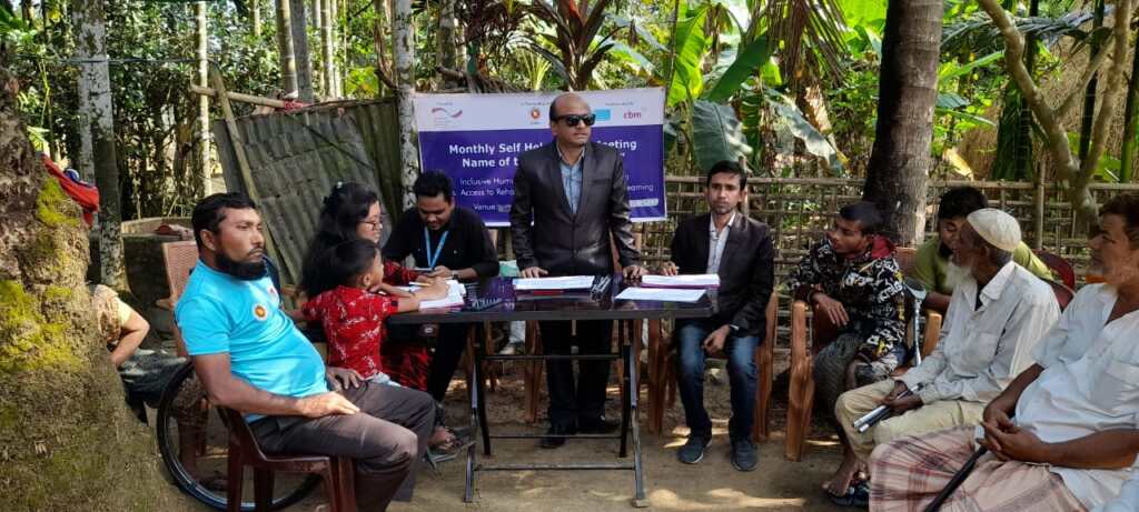 A community meeting taking place outdoors in a rural area of Bangladesh, with men and women seated around a table under trees and surrounded by greenery. A man in a black suit and sunglasses stands at the centre, addressing the group. A banner behind the table reads “Monthly Self Help Group Meeting” and includes logos of CBM Global, the Government of Bangladesh, and partners, highlighting inclusive humanitarian action and access to rehabilitation services.