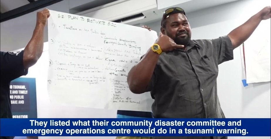 n Fiji, a man gestures while presenting during a community disaster preparedness session. Behind him, another participant holds a large sheet of paper listing plans to reactivate the emergency operations center during a tsunami warning. A caption at the bottom explains that the group discussed what their community disaster committee would do in such an event.