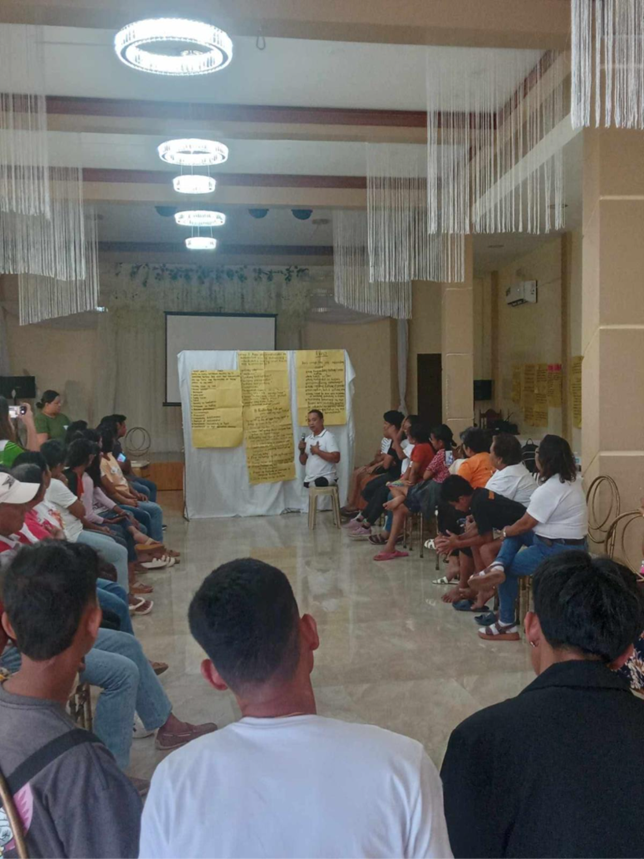 A community workshop in the Philippines where participants sit in a large circle listening to a facilitator speaking in front. Large sheets of yellow paper with handwritten notes are displayed on the wall behind the facilitator. The session is held indoors under bright ceiling lights.