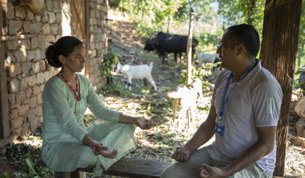 A CMC Nepal staff member facilitates a calming breathing exercise with a woman in Nepal as part of a community-based mental health and psychosocial support session. They sit outside a rural home, surrounded by trees and livestock, focusing on mindfulness and emotional wellbeing.