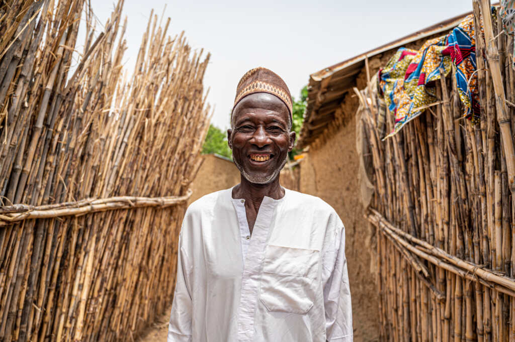 An older man stands smiling in a narrow pathway in a rural community in Nigeria, surrounded by tall woven fences made of sticks. He wears a light-coloured traditional shirt and a patterned cap
