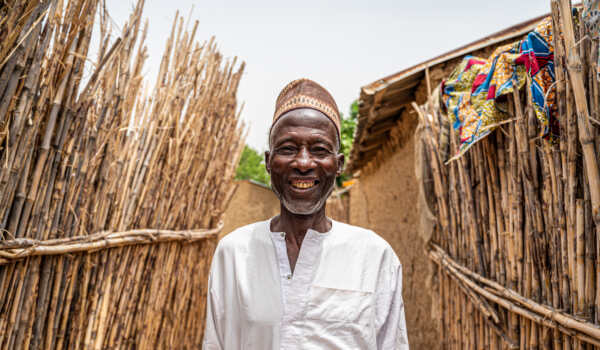 An older man stands smiling in a narrow pathway in a rural community in Nigeria, surrounded by tall woven fences made of sticks. He wears a light-coloured traditional shirt and a patterned cap
