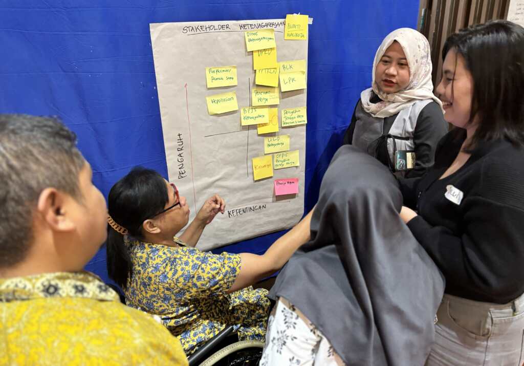 A group of people gather around a large sheet of paper taped to a wall, displaying a stakeholder mapping chart with handwritten labels and colorful sticky notes arranged in columns. One participant, seated in a wheelchair, gestures toward the chart while others stand close by, looking at the notes and discussing the content.