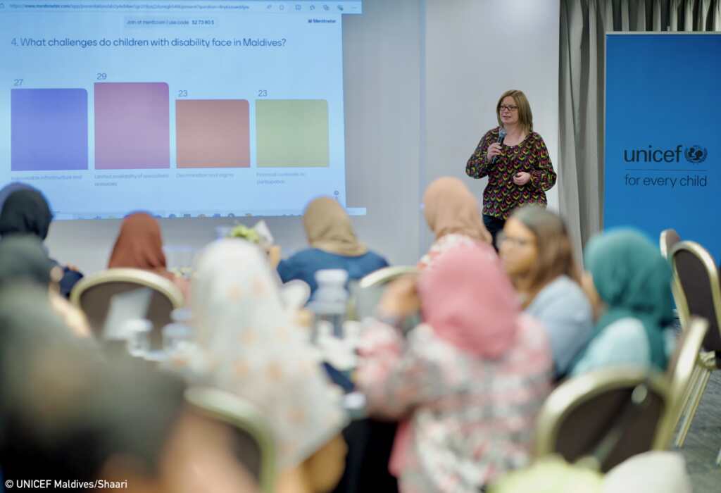 A woman speaks into a microphone during a workshop in the Maldives while participants listen from round tables. A presentation screen behind her shows survey results about the challenges faced by children with disabilities, and a UNICEF banner stands to the side of the room.