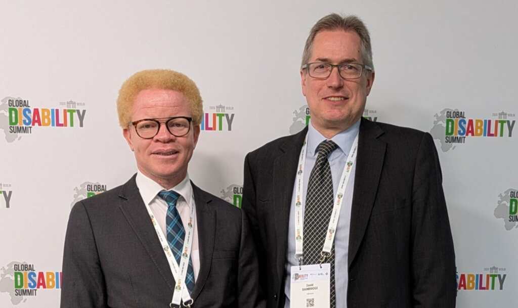 Two men in suits stand side by side in front of a Global Disability Summit 2025 backdrop in Berlin, smiling for a photo.