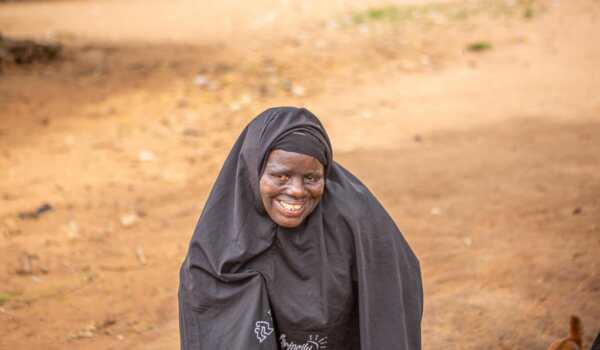 Fatima, a woman from Nigeria wearing a black hijab, smiles while standing outdoors on a sandy path.