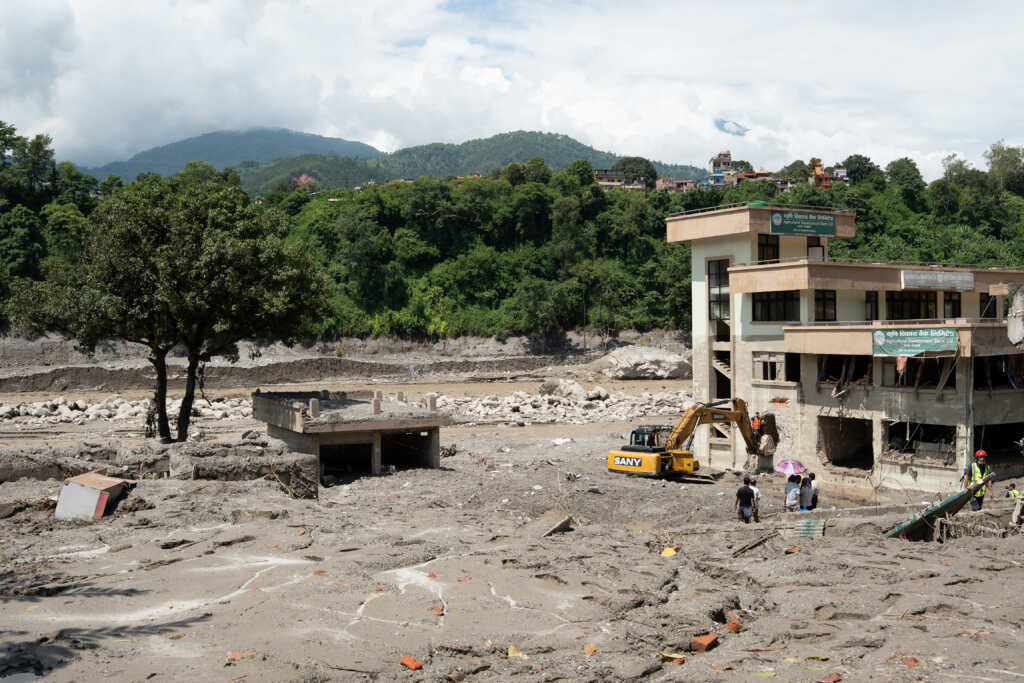 People and an excavator beside a damaged building surrounded by mud, rocks and debris after flooding in Nuwakot, Nepal.