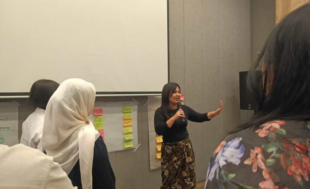 A woman speaks into a microphone and gestures to participants during a training session in Indonesia. Colourful sticky notes are displayed on the wall behind her.